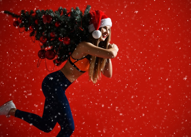 Santa hat Christmas sporting woman holding xmas tree on her shoulders. Winner energy he red background. it's snowing